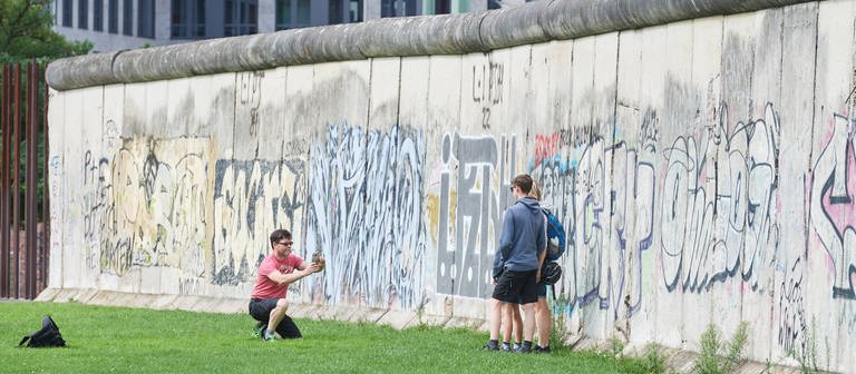 Menschen machen Fotos an Resten der Berliner Mauer. Die Teilung Deutschlands hat das Leben vieler in Ost und West geprägt. (Foto: IMAGO / epd)