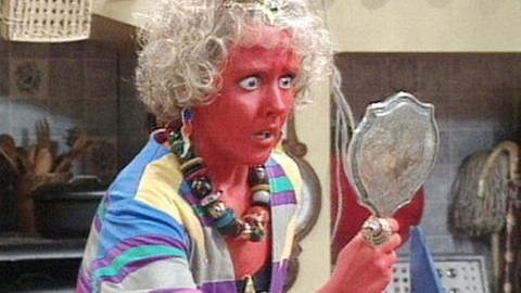 Eine Frau mit roter Haut und in bunten Klamotten schaut entsetzt in einen Handspiegel. (Foto: SWR - Screenshot aus der Sendung)
