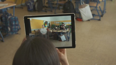 Auf einem Tablet sieht eine Frau einen Klassenraum und die Holografie einer Zeitzeugin