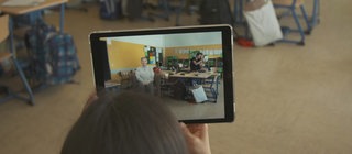 Auf einem Tablet sieht eine Frau einen Klassenraum und die Holografie einer Zeitzeugin