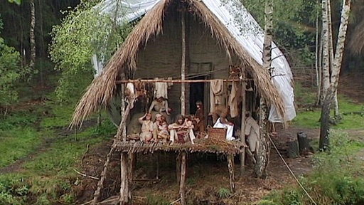 Sippe sitzt auf dem Vorbau ihres Pfahlhauses.
