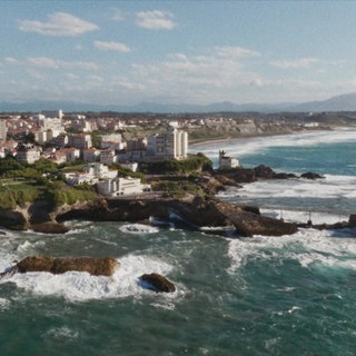 Felsige Küstenlandschaft in Biarritz mit Meer, Strand, Uferbebauung und Bergen im Hintergrund