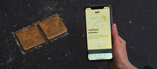 Eine Hand mit Smartphone, auf dem die App "Stolpersteine" zu sehen ist