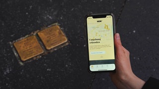 Eine Hand mit Smartphone, auf dem die App "Stolpersteine" zu sehen ist