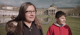 Ein Junge und ein Mädchen stehen auf einer Wiese vor einem großen Gebäude.