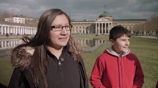 Ein Junge und ein Mädchen stehen auf einer Wiese vor einem großen Gebäude.