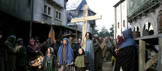 Kinder in mittelalterlicher Kleidung ziehen durch ein Dorf und tragen ein Kreuz.
