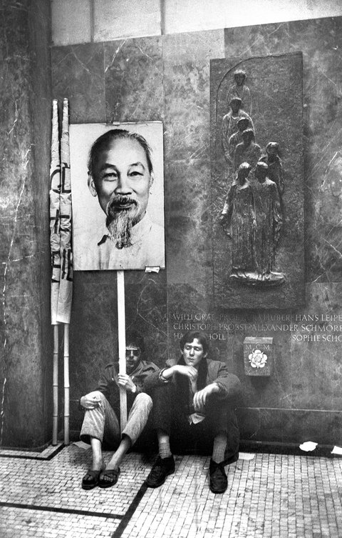 Zwei junge Männer sitzen mit Ho-Chi-Minh-Transparent vor einer Wand