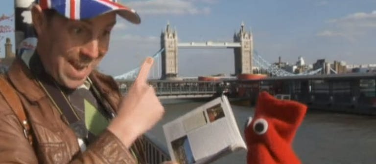 Ein Mann trägt eine Kappe, die mit dem Unionjack bedruckt ist. Neben ihm eine rote Strumpfhandpuppe. Sie stehen vor der Tower Bridge in London. (Foto: WDR - Screenshot aus der Sendung)