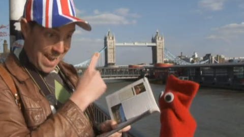 Ein Mann trägt eine Kappe, die mit dem Unionjack bedruckt ist. Neben ihm eine rote Strumpfhandpuppe. Sie stehen vor der Tower Bridge in London. (Foto: WDR - Screenshot aus der Sendung)