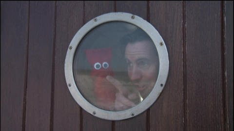 Ein Mann und eine rote Strumpfhandpuppe sind auf einem Schiff und schauen durch ein Bullauge. (Foto: WDR - Screenshot aus der Sendung)