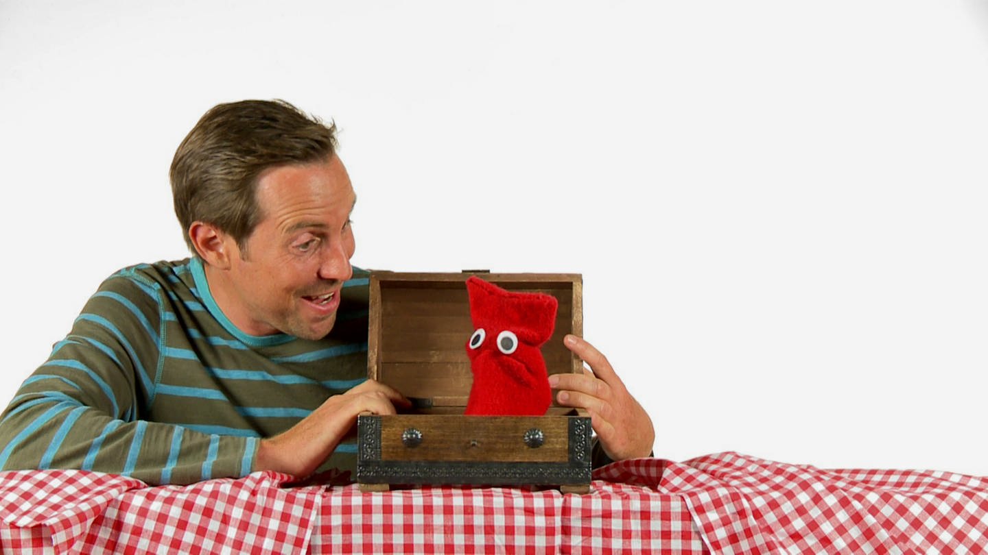 Ein Mann findet eine rote Strumpfhandpuppe in einer Holzkiste. (Foto: WDR - Screenshot aus der Sendung)