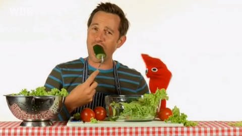 Ein Mann beißt in eine Gurke, neben ihm eine rote Strumpfhandpuppe, vor ihnen auf dem Tisch liegt Salat.  (Foto: WDR - Screenshot aus der Sendung)