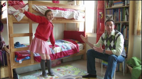 Ein Mädchen steht vor ihrem Hochbett und trägt eine Schuluniform, ein Mann sitzt neben ihr auf einem Stuhl. (Foto: WDR - Screenshot aus der Sendung)