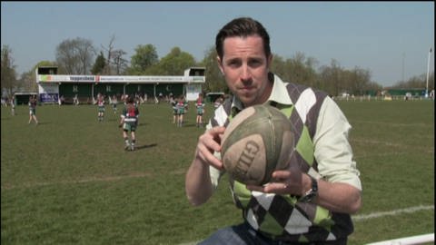 Ein Mann steht vor einem Sportplatz und hält einen Rugbyball in der Hand. Hinter ihm spielen zwei Mannschaften gegeneinander Rugby. (Foto: WDR - Screenshot aus der Sendung)