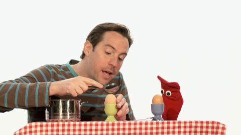 Ein Mann öffnet mit einem Löffel ein gekochtes Ei. Eine rote Strumpfhandpuppe schaut ihm dabei zu.  (Foto: WDR - Screenshot aus der Sendung)