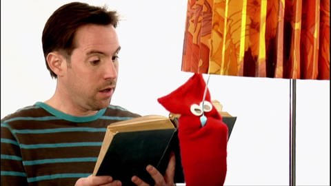 Ein Mann liest ein Buch, eine rote Strumpfhandpuppe macht eine Stehlampe neben ihm aus. (Foto: WDR - Screenshot aus der Sendung)