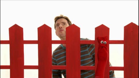 Ein Mann sucht eine rote Strumpfhandpuppe. Diese versteckt sich hinter einem roten Zaun. (Foto: WDR - Screenshot aus der Sendung)