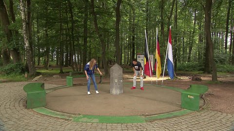 Ein Mann und eine Frau stehen im Wald um einen kleinen Steinobelisk. Im Hintergrund wehen die Fahnen von Deutschland, Belgien und den Niederlanden. (Foto: WDR - Screenshot aus der Sendung)