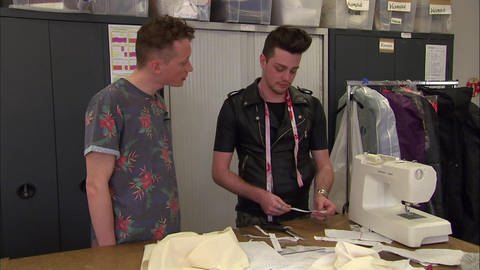 Zwei junge Männer in einer Werkstatt. Einer der beiden trägt ein Maßband um den Hals und arbeitet an einer Nähmaschine. (Foto: WDR - Screenshot aus der Sendung)