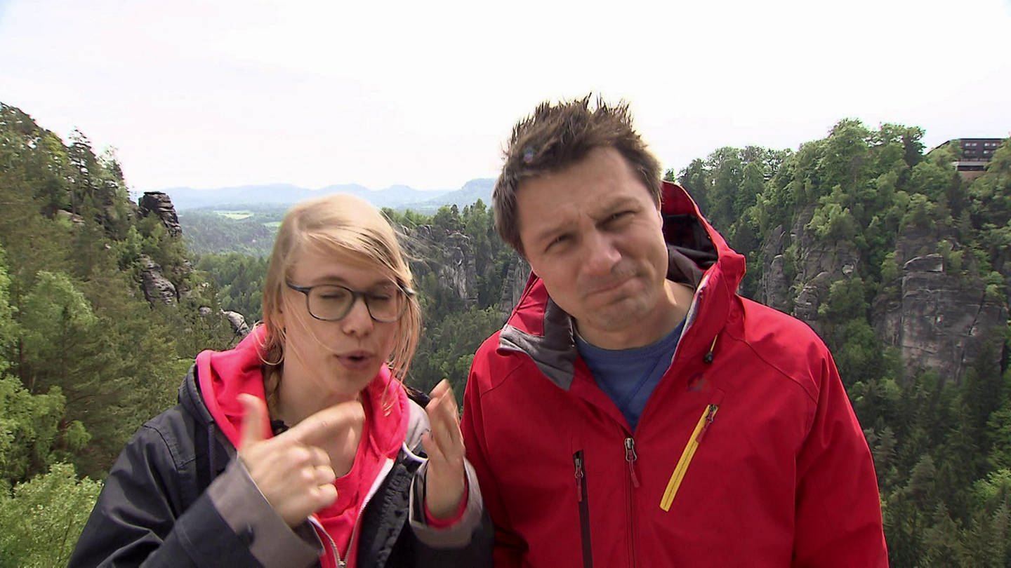 Eine Frau und ein Mann stehen in einem Gebirge, umgeben von Wäldern. (Foto: SWR / BR / WDR - Screenshot aus der Sendung)