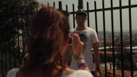 Ein junger Mann steht vor einem Zaun, hinter ihm liegt die Stadt. Eine Frau macht ein Foto von ihm. (Foto: WDR - Screenshot aus der Sendung)