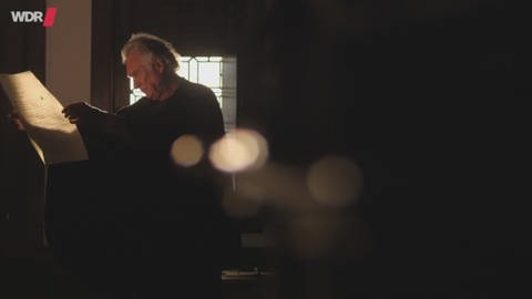 Ein Mann steht in einem dunklen Raum und studiert ein großes Stück Papier.  (Foto: WDR - Screenshot aus der Sendung)