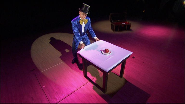 Ein Zauberer steht an einem gedeckten Tisch und zieht an der Tischdecke. (Foto: SWR - Screenshot)