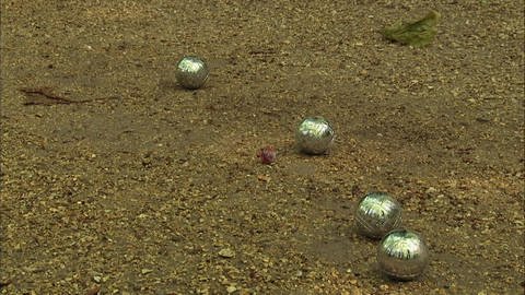 Vier silberne Boulekugeln und eine kleiner roter Ball liegen auf sandigem Boden.  (Foto: WDR - Screenshot aus der Sendung)