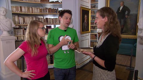 Drei Personen stehen in einer Bibliothek. Hinter ihnen eine Marmorbüste und mehrere Bilder. (Foto: SWR / BR / WDR - Screenshot aus der Sendung)