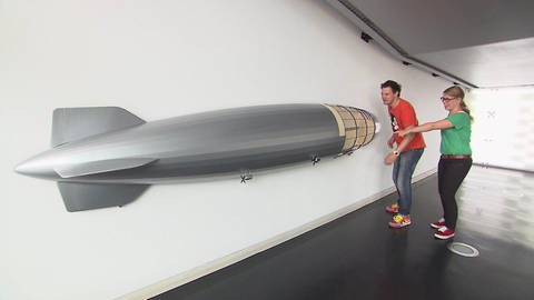 Ein Mann und eine Frau stehen in einer Halle vor dem Modell eines Zeppelins.  (Foto: SWR / BR / WDR - Screenshot aus der Sendung)
