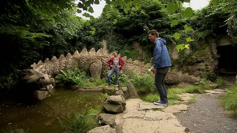 Ein Mann und eine Frau stehen in einem Garten. Vor ihnen liegt eine große Drachenstatue aus Stein.  (Foto: WDR - Screenshot aus der Sendung)