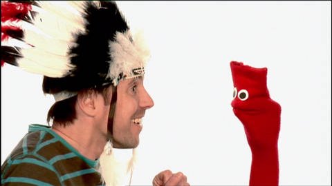 Ein Mann trägt indigenen Federschmuck, eine rote Strumpfhandpuppe schaut ihm dabei zu. (Foto: WDR - Screenshot aus der Sendung)