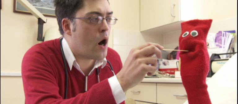 Ein Arzt hält einer roten Strumpfhandpuppe einen Holzstab in den geöffneten Mund. (Foto: WDR - Screenshot aus der Sendung)