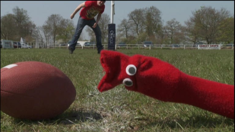 Eine rote Strumpfhandpuppe liegt auf dem Rasen, neben ihr ein Rugbyball. (Foto: WDR - Screenshot aus der Sendung)