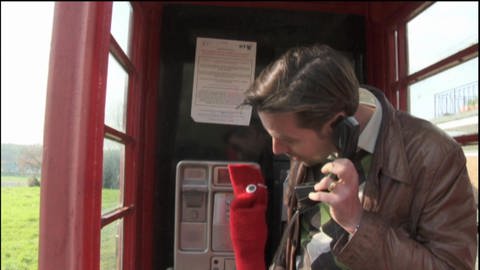 Ein Mann und eine rote Strumpfhandpuppe telefonieren in einer Telefonzelle. (Foto: WDR - Screenshot aus der Sendung)