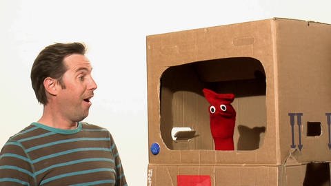 Eine rote Strumpfhandpuppe sitzt in einem Fernseher aus Pappe. Ein Mann schaut ihr dabei zu. (Foto: WDR - Screenshot aus der Sendung)