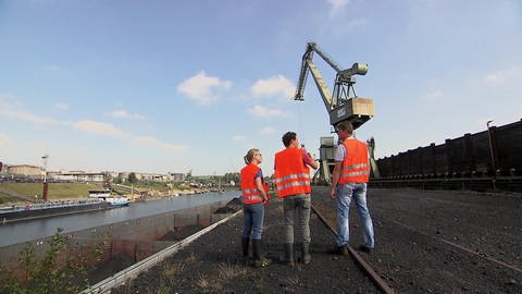 Drei Menschen stehen in orangenen Warnwesten am Duisburger Hafen. Im Hintergrund steht ein großer Kran und entlädt einen Container. (Foto: WDR - Screenshot aus der Sendung)