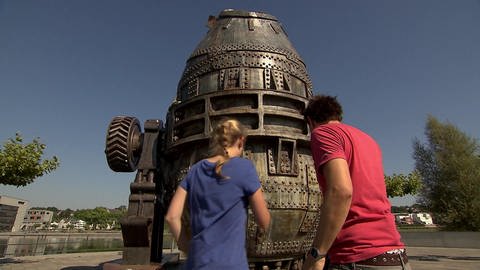 Ein Mann und eine Frau stehen vor einem 7 Meter großen, birnenförmigen Stahlkochtopf am Phönix See. (Foto: WDR - Screenshot aus der Sendung)