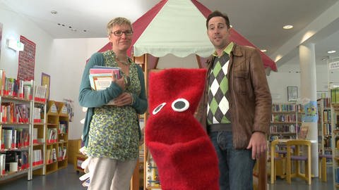 Ein Mann und eine Frau stehen in einer Bibliothek. Vor ihnen ist eine rote Strumpfhandpuppe. (Foto: WDR - Screenshot aus der Sendung)