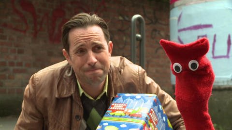 Ein Mann und eine rote Strumpfhandpuppe sitzen auf dem Bürgersteig. Der Mann trägt ein Geschenk. (Foto: WDR - Screenshot aus der Sendung)