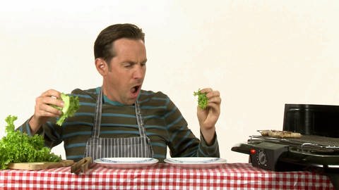 Ein Mann sitzt an einem Tisch. Er macht Salat und brät Würstchen.  (Foto: WDR - Screenshot aus der Sendung)