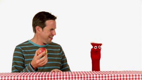 Ein Mann hält einen Apfel in der Hand und schaut dabei bestürzt eine rote Strumpfhandpuppe an.  (Foto: WDR - Screenshot aus der Sendung)