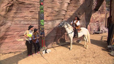 Ein Mann und eine Frau stehen als Cowboys verkleidet an einem Marterpfahl. Neben ihnen sitzt eine Frau auf einem Pferd. (Foto: SWR / BR / WDR - Screenshot aus der Sendung)