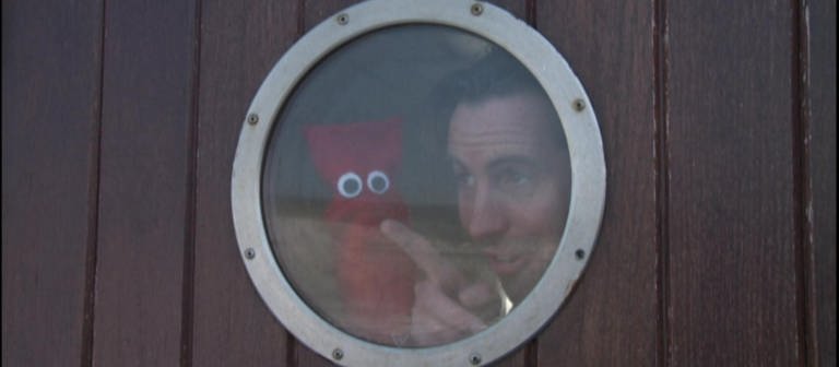 Ein Mann und eine rote Strumpfhandpuppe sind auf einem Schiff und schauen durch ein Bullauge. (Foto: WDR - Screenshot aus der Sendung)