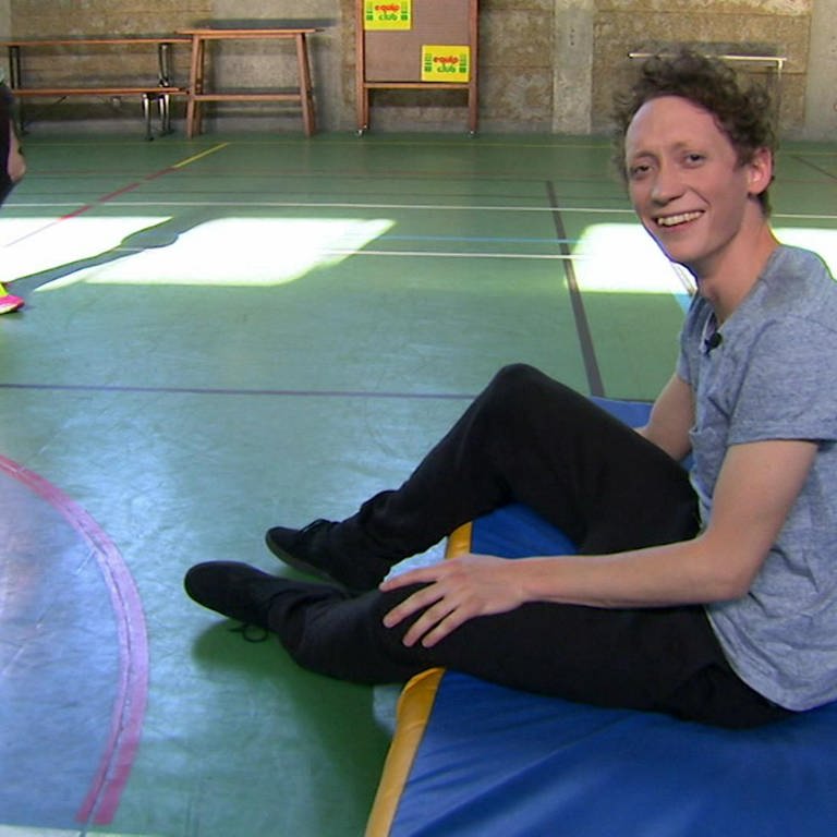 Ein junger Mann sitzt lachend auf einer Matte in einer Sporthalle. (Foto: WDR - Screenshot aus der Sendung)