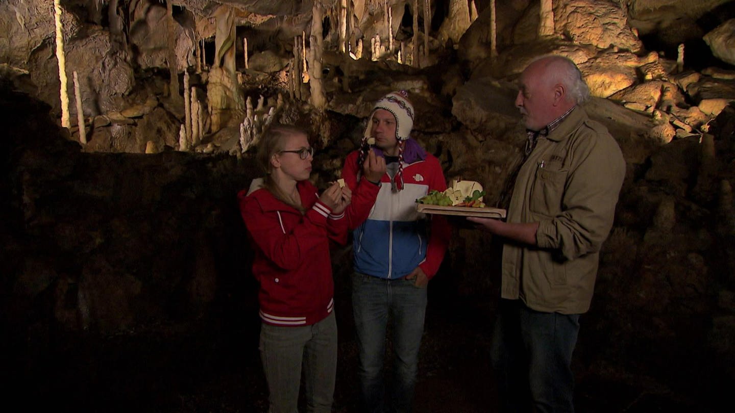 Ein Mann und eine Frau stehen in einer Tropfsteinhöhle und essen Käse. Ein älterer Mann hält ihnen eine Käseplatte hin. (Foto: WDR - Screenshot aus der Sendung)