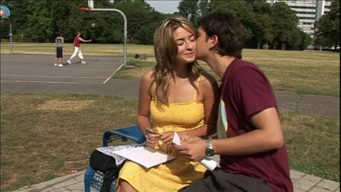 Ein junges Pärchen sitzt neben einem Basketballplatz, der junge Mann küsst die junge Frau auf die Wange. (Foto: WDR)