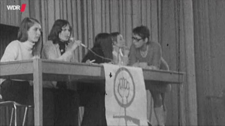 Schwarz-weiß Bild von einem langen Tisch auf einer Tribüne. An dem Tisch sitzen fünf Frauen, eine spricht in ein Mikrofon. Es hängt eine Fahne mit geballter Faust vor dem Tisch. (Foto: WDR)