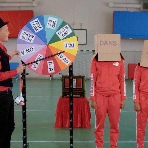Ein junger Mann steht an einem bunten Glücksrad, hinter ihm zwei Personen mit Kartons auf dem Kopf. Auf einem Karton steht "Dans", auf dem anderen "É". (Foto: WDR)